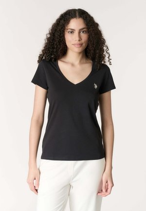 Donna con capelli ricci che indossa una maglietta nera a maniche corte con scollo a V e pantaloni bianchi, in piedi davanti a uno sfondo semplice.