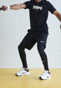 Camiseta negra de Nike con estampado "HWPO", pantalones cortos negros y leggings negros. Zapatillas Nike blancas y rojas. Suelo de color claro y fondo de ladrillo blanco.