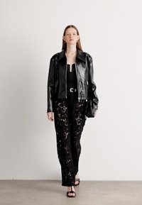 Giacca in pelle nera con finitura texturizzata, body nero aderente e pantaloni in pizzo nero trasparente. Gli accessori includono una piccola borsa nera.