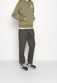Olivgrön huvtröja med ficka fram och vit logotyp, parat med mörkgröna cargobyxor och marinblå sneakers med vita detaljer.