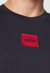 Námořnické tričko s kulatým výstřihem, s červenou obdélníkovou vyšívanou nášivkou, na které je napsáno "HUGO" černě, vyrobené z měkké, elastické tkaniny.