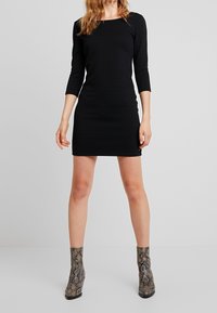 Musta, ribattu pitkähihainen mekko neliönmuotoisella kaula-aukolla. Yhdistetty käärmeennahkakuosisilla nilkkabootseilla. Mekko ulottuu reiteen puoliväliin.