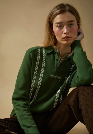 Joven con cabello largo que lleva un suéter verde con patrones a rayas y pantalones marrón oscuro, apoyando la cabeza en la mano sobre un fondo beige.