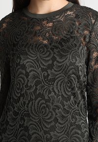 Mörkgrön spetsblus med långa ärmar och blommönster, med en enfärgad rund halsringning och texturerat överdrag. Genomskinliga sektioner är synliga.