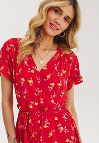 Czerwona sukienka w kwiaty z krótkimi falbaniastymi rękawami, zapinana na guziki z przodu, dekoltem w kształcie litery V oraz związanym w talii pasem. Charakteryzuje się żółtymi i różowymi wzorami kwiatowymi. Wykonana z miękkiego materiału.