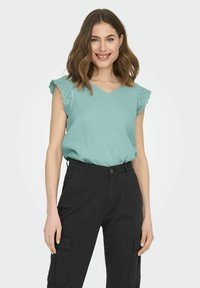 Top verde menta con scollo a V e maniche in pizzo decorativo a forma di conchiglia, abbinato a pantaloni cargo neri a vita alta. Tessuto morbido e leggero.