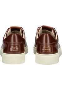 Zapatillas de piel marrón con cuello acolchado, suela de goma blanca, detalle del logotipo dorado en el talón y textura suave. Diseño moderno y de perfil bajo.