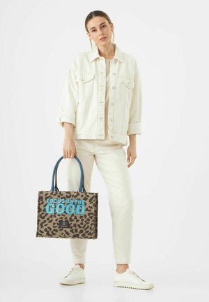 Leopardenmuster-Canvas-Tragetasche mit blauer Schrift "FOKUSSIERE AUF DAS GUTE", stabilen Trägern und rechteckiger Form. Modells trägt ein helles Outfit.