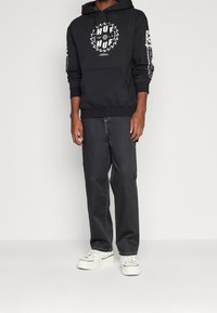 Czarna bluza z kapturem z białym graficznym logo, kangurkowa kieszeń, noszona z ciemnymi, luźnymi spodniami i białymi sneakersami. Gładka bawełniana tekstura, casualowy design.