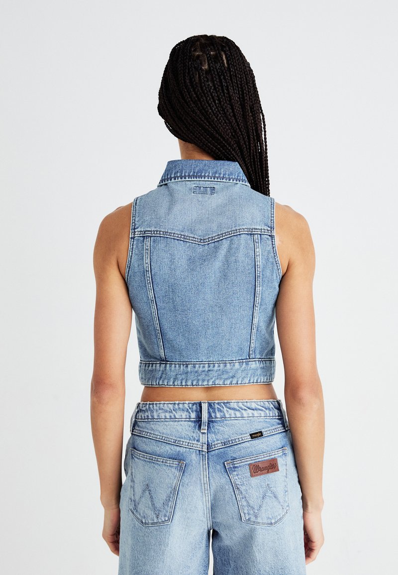 Gecroppte Jeansweste in Hellblau mit klassischem Kragen, ärmellosem Design und sichtbaren Nähdetails. Kombiniert mit hoch taillierten Jeans.