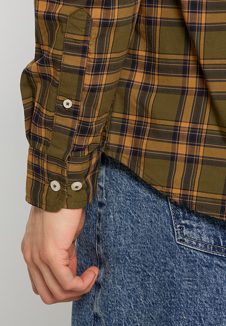 Una mano maschile appoggiata accanto alla tasca di un paio di jeans blu, che indossa una camicia a maniche lunghe a quadri marrone e blu con bottoni bianchi sui polsini.