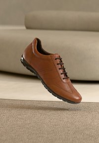 Baskets en cuir marron avec lacets et semelle noirs, légèrement surélevées au-dessus d’un tapis beige et d’un canapé moderne clair en arrière-plan.