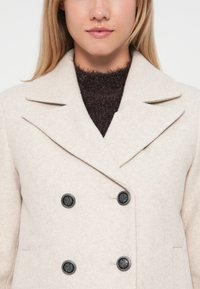 Manteau beige croisé en mélange de laine avec des boutons noirs, larges revers et col texturé, porté sur un pull en tricot marron foncé.