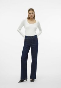 Vit långärmad åtsittande topp kombinerad med mörkblå högmidjade utsvängda jeans, accentuerad med ljusa sömmar och spetsiga svarta skor.