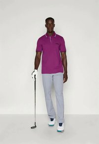 Vijolična polo majica s kontrastnim ovratnikom in logotipom, v kombinaciji s svetlo sivimi hlačami. Model drži golf palico in nosi bele in modre čevlje.