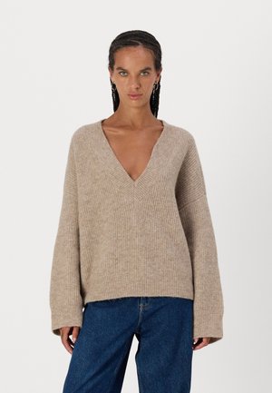 Beige gestrickter Pullover mit tiefem V-Ausschnitt, weiten Ärmeln und geripptem Muster. Kombiniert mit hoch taillierten blauen Jeans.