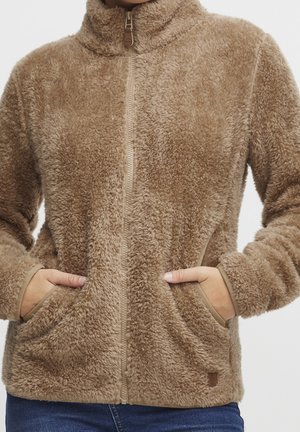 Veste polaire beige zippée avec col montant et poches latérales, portée avec les mains dans les poches, montrée du cou aux hanches.
