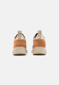 Die Sneakers bestehen aus einem hellbraunen Wildledermaterial mit goldenen Akzenten, haben eine runde Zehenpartie, einen gepolsterten Kragen und eine strukturierte beigefarbene Sohle. Kurzer Ziehriemen hinten.