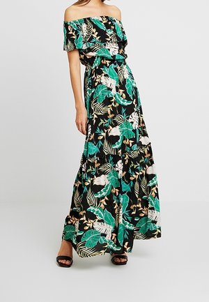 Vrouw draagt een zwarte maxi-jurk met off-shoulder en tropische bladprint in groen en wit, gecombineerd met zwarte open-toe hakken.