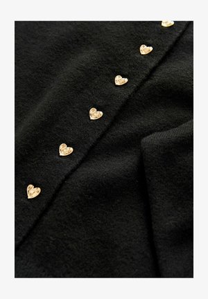 Musta neule, jossa on rivi kultaisia sydämenmuotoisia nappeja edessä, valmistettu pehmeästä, teksturoidusta kankaasta.