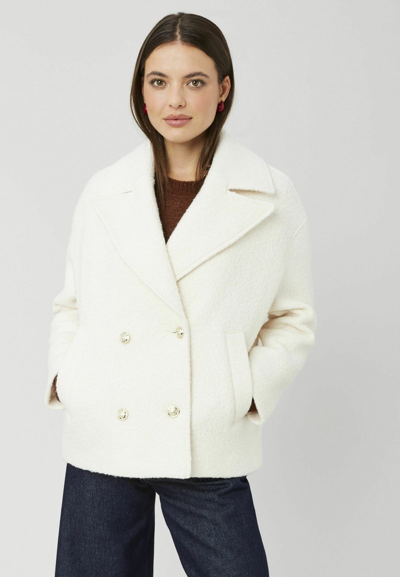 Manteau croisé crème avec grands revers, en tissu texturé. Comprend deux poches avant et des boutons dorés, associé à un jean foncé.