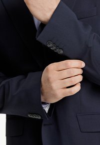 Granatowa marynarka o gładkiej fakturze, z mankietami na cztery guziki i jednym przednim kieszeniom. Pod spodem częściowo widoczna jest koszula w paski.