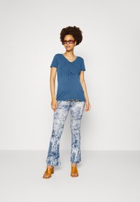 Camiseta azul de mangas curtas com detalhe de nó na cintura, combinada com calças flare em tie-dye azul e branco, e sandálias de salto bloco em tom bege.