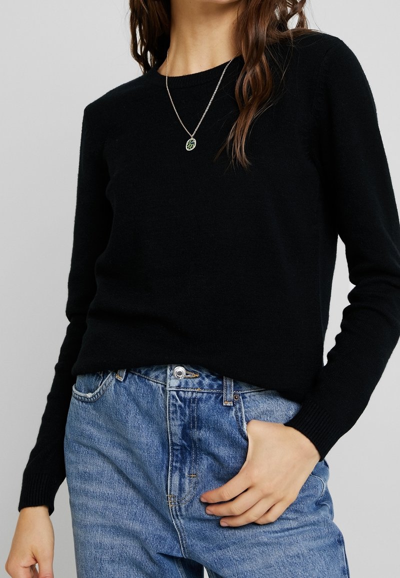 Femme portant un pull à manches longues noir, un jean en denim bleu et un collier en argent avec un pendentif circulaire vert.