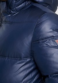 Námořnický modrý prošívaný kabát s lesklou, hladkou texturou. Má kapuci a viditelné švy. Obsahuje logovou nášivku na rameni.