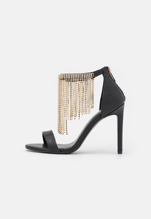 Sort højhælet sandal med et elegant design, med en frynsekant af guldkæder. Glatte læderremme og en lynlåslukning bagpå.