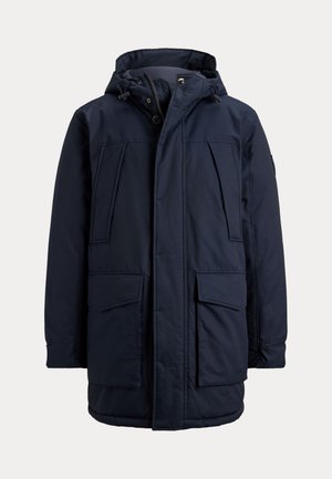 Homme portant un manteau d'hiver à capuche bleu marine par-dessus un pull bleu, une chemise bleu clair à col, un pantalon gris et des chaussures marron sur un fond uni.