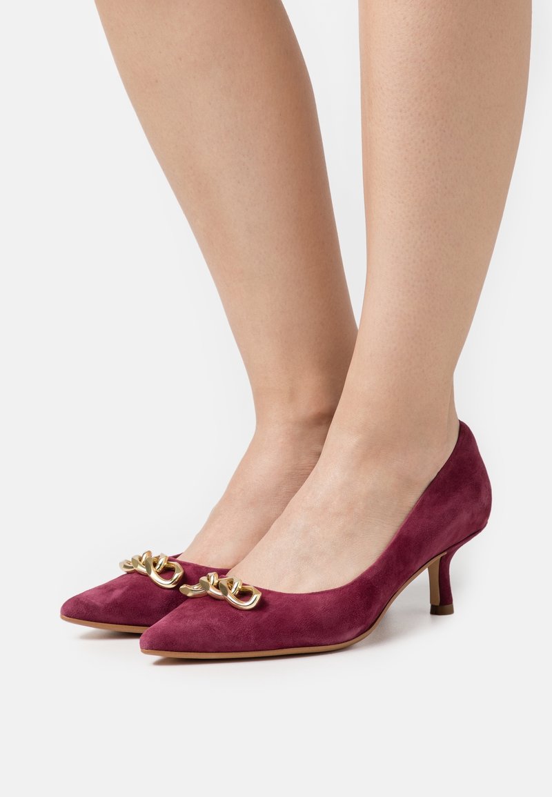 Femme portant des escarpins en daim bordeaux à talons kitten, bouts pointus et détails de chaîne dorée à l'avant.