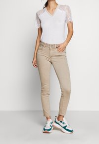 Camiseta blanca de cuello en V con mangas de encaje, jeans ajustados beige y zapatillas multicolores con acentos en azul verdoso, blanco y rosa.