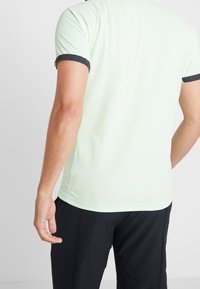 T-shirt vert clair à manches courtes avec une finition texturée et des poignets bordés de marine foncé, associé à un pantalon noir. Vue de dos.