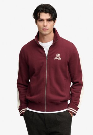 Jonge man met een bordeauxrode jas met rits, met witte strepen op de mouwen en een wit logo op de borst, staand met handen in de zakken.