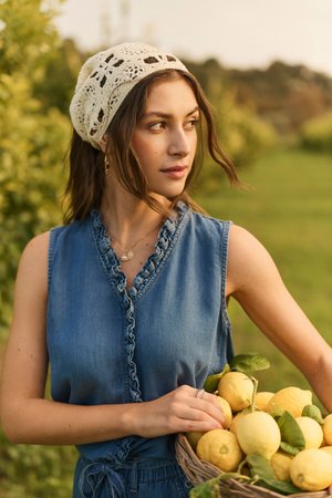 Nő horgolt fejkendőben és farmer felsőben, citromokkal teli kosarat tart egy napsütötte gyümölcsösben, oldalra néz.