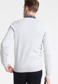 Šedý svetr s kulatým výstřihem, elastickými manžetami a spodním lemem, přičemž je viditelný límec z modré kostkované košile. Hladký povrch tkaniny.