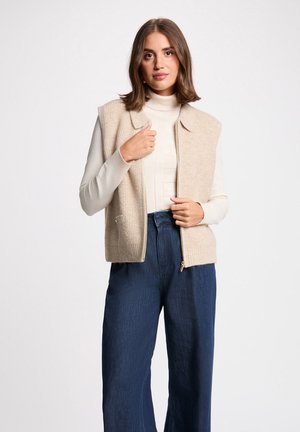 Vrouw die naar voren kijkt, draagt een beige vest met rits over een crèmekleurige coltrui en wijde blauwe broek tegen een effen lichte achtergrond.
