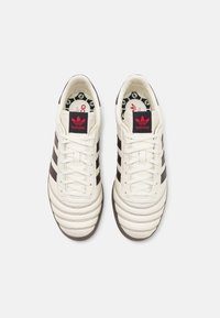 Vita lädersneakers med tre mörka ränder, gummisula och röd Adidas-logga på tungan. Detaljerna inkluderar strukturerad sömnad.