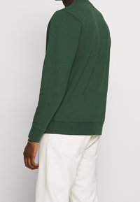 Uomo con camicia a maniche lunghe verde scuro e pantaloni bianchi, rivolto di lato su uno sfondo chiaro e uniforme, braccialetto d'argento al polso sinistro.