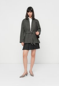Belted black and white checked jacket with a collar, long sleeves, and button closure, paired with a pleated black skirt and grey shoes.