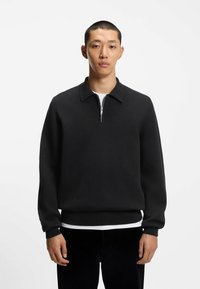 Pullover nero con colletto a zip realizzato in un tessuto testurizzato. Maniche e orlo a coste, indossato sopra una camicia bianca. Design semplice e minimal.