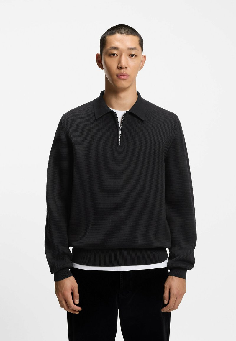 Pullover nero con colletto a zip realizzato in un tessuto testurizzato. Maniche e orlo a coste, indossato sopra una camicia bianca. Design semplice e minimal.