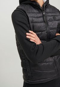 Gilet piumino nero con design trapuntato, chiusura frontale con zip, abbinato a un hoodie nero a maniche lunghe. Presenta maniche aderenti e scollatura rotonda.