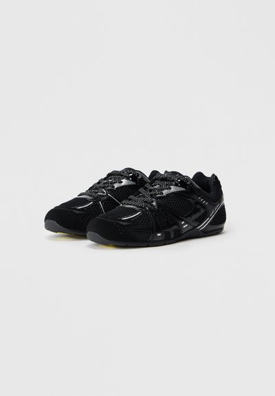 Zapatillas deportivas negras con detalles de malla y brillantes, cordones con patrón y suela de perfil bajo sobre un fondo gris claro.