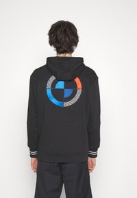 Felpa nera con cappuccio caratterizzata da un design ricamato multicolore a forma di cerchio sul retro, con dettagli a scacchi e polsini a righe. Il materiale sembra morbido.