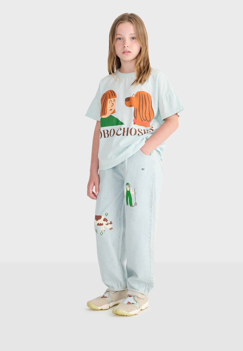 Młoda dziewczynka w jasnoniebieskiej koszulce z grafiką kreskówkowego psa i dziewczynki oraz jasnych dżinsach z naszywkami rolnika i krowy, stojąca z jedną ręką w kieszeni.