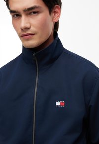 Młody mężczyzna w granatowej kurtce zapinanej na zamek z logo Tommy Jeans na piersi, patrzący lekko w bok na białym tle.