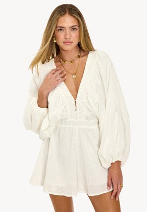 Femme aux longs cheveux blonds portant une mini robe blanche à œillets avec des manches bouffantes et des colliers superposés en or en forme de coquillage sur un fond blanc.