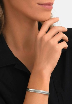 Femme aux lèvres neutres et chemise noire touchant son menton, portant un fin bracelet en argent au poignet.